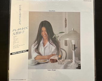 Può includere: Una copertina di un album in vinile bianco con una donna con lunghi capelli scuri seduta a un tavolo con una lampada e una pianta. L'album è intitolato "Grey Skies" di Taeko Ohnuki. Il testo "White Color Vinyl" è stampato sulla copertina.