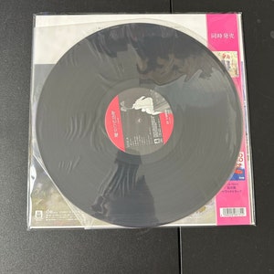 K&ouml;nnte beinhalten: Eine schwarze Schallplatte mit einem roten Etikett, auf dem "魔女の宅急便" (Majo no Takkyubin) und "SIDE-A" steht. Die Schallplatte befindet sich in einer durchsichtigen Plastikt&uuml;te. Die R&uuml;ckseite der H&uuml;lle hat einen rosa Hintergrund mit japanischem Text und den Worten "同時発売" (Douji Hatsuba) und "紅の部" (Kurenai no Bu).