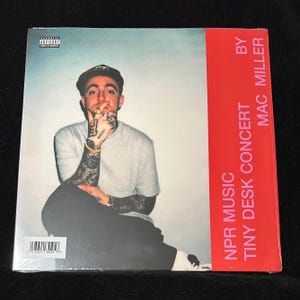 May include: Album cover featuring Mac Miller, seated and smoking, with tattoos visible on his arms and hands. The album cover has a red vertical stripe with the text "NPR Music Tiny Desk Concert by Mac Miller."