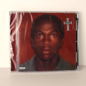 May include: A CD case featuring a portrait of a person with a dark complexion against a red background. The CD case has a sticker that reads "SON OF SPERGY" in white text. The case is sealed in clear plastic.