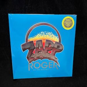 May include: A sealed vinyl record album with a bright blue cover. The album features the band name "ZAPP" in a stylized, graffiti-style font, with "ROGER" and "ALL THE GREATEST HITS" in gold lettering. A circular sticker is in the upper right corner.