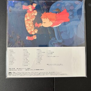 May include: A vinyl record album cover featuring a colourful illustration of a young girl with red hair and a red dress, swimming with a large, blue, jellyfish-like creature. The album is titled "Ponyo" and features the names of the composer, conductor, and singer. The album cover also includes a track list for each side of the record.