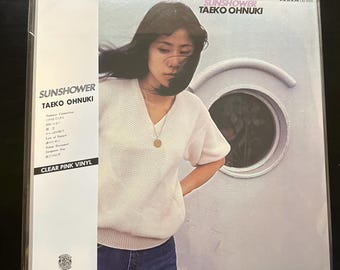 Può includere: Una copertina di un album in vinile con una donna che indossa un maglione bianco e jeans blu. L'album è intitolato "SUNSHOWER" di "TAEKO OHNUKI" ed è sull'etichetta "PANAM". L'album è un vinile rosa chiaro.