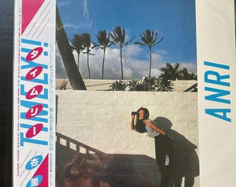 Può includere: Una copertina di un album in vinile con una donna in camicia a righe e gonna nera in piedi davanti a un muro bianco. Lo sfondo mostra palme e un cielo blu. Il titolo dell'album è "Timely" e il nome dell'artista è "ANRI".