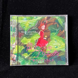 May include: A CD case featuring the animated film "The Secret World of Arrietty" soundtrack. The cover art depicts a girl in a red dress amidst green foliage. Japanese text is visible on the cover.