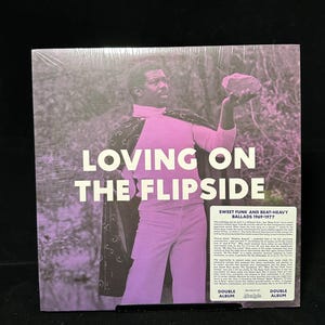 May include: A vinyl record album titled "Loving on the Flipside" with a purple and white design. The album cover features a man in a pink outfit holding a rock. Text on the album reads "Sweet Funk and Beat-Heavy Ballads 1969-1977."