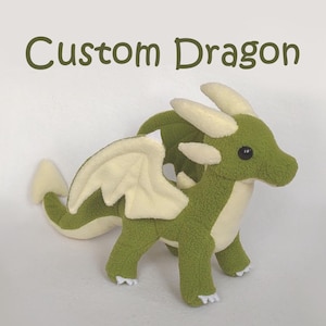 May include: A plush green dragon with cream-colored wings, horns, and belly. The stuffed animal has black button eyes and is labeled "Custom Dragon" in green text.