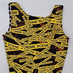 May include: A sleeveless one-piece swimsuit featuring a black background with a repeating pattern of yellow crime scene tape. The tape is printed with the text CRIME SCENE in black capital letters, interspersed with red splatters.