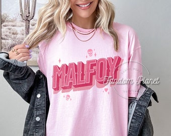 Puede incluir: Camiseta rosa de gran tamaño con un gráfico rosa y rojo de la palabra "MALFOY" en un estilo retro con calaveras y corazones.