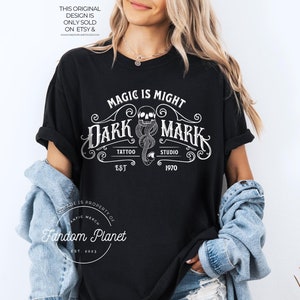 May include: Black t-shirt with a white graphic design that says "Magic is Might Dark Mark Tattoo Studio Est 1970". The graphic features a skull with a snake wrapped around it.