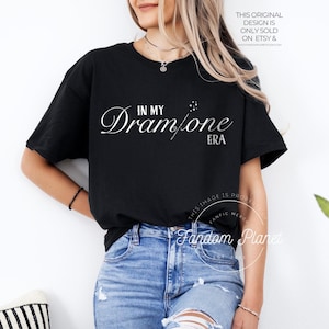 May include: A black t-shirt with the text "In My Dram/one Era" printed in white on the front. The shirt is being worn by a person with blonde hair and blue jeans.