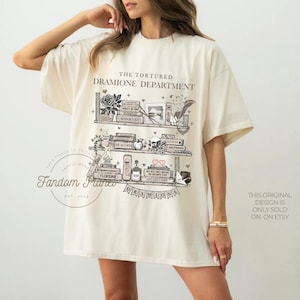 May include: A beige t-shirt with a graphic design of a bookshelf with books, a skull, and a potion bottle. The text "The Tortured Dramione Department" is printed above the bookshelf. The text "Dramione" is printed below the bookshelf.