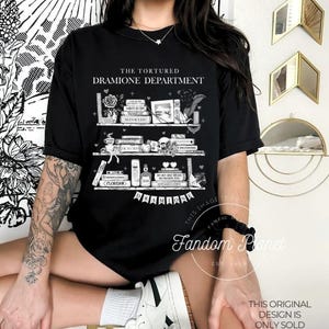 May include: Black t-shirt with a white graphic of a bookshelf with books and other objects. The text "The Tortured Dramione Department" is printed on the shirt.
