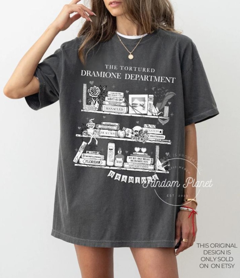 May include: A dark gray t-shirt with a white graphic of a bookshelf with books, flowers, and other decorative items. The text "The Tortured Dramione Department" is printed above the bookshelf. The text "Fandom Planet Est 2021" is printed below the bookshelf.