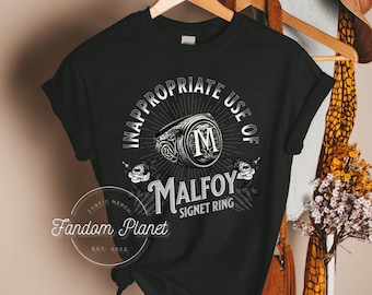 Puede incluir: Camiseta negra con un diseño gráfico blanco que dice "Uso inapropiado del anillo de sello de Malfoy". El gráfico presenta un anillo estilizado con una "M" en el centro.