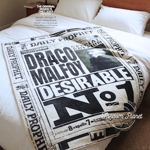 May include: A white throw blanket featuring a black graphic print of a Daily Prophet newspaper front page. The design includes the text Draco Malfoy, Desirable No. 1, and a photographic image of a person. The border repeats the Daily Prophet name.