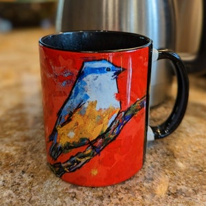 May include: A red coffee mug with a black handle and a colorful painting of a bluebird on a branch. The bird is painted in shades of blue, yellow, and orange.