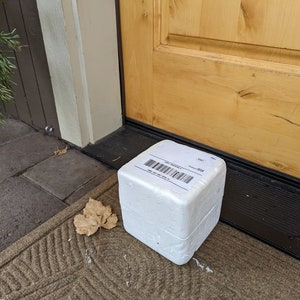 Op de afbeelding: Een witte styropor doos met een verzendlabel op de bovenkant. Het label heeft een barcode en de tekst "ORDER TRACKING" en "ORDER #". De doos staat op een bruine deurmat.
