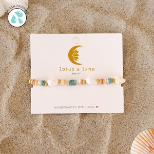 Könnte beinhalten: Fußkettchen mit weißen, beigen, goldenen und türkisfarbenen Perlen. Das Fußkettchen wird auf einer weißen Karte mit dem Text "lotus & luna ANKLET" und "HANDCRAFTED WITH LOVE" präsentiert. Der Hintergrund ist Sand.