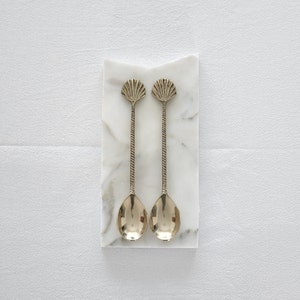 May include: Two silver-coloured spoons with seashell-shaped tops and twisted handles rest on a white marble rectangular base. The spoons have oval-shaped bowls. The marble base has a natural veining pattern.