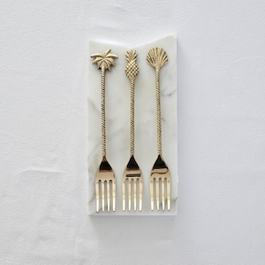 Peut inclure: Trois fourchettes à cocktail en métal doré avec des motifs tropicaux différents : un palmier, un ananas et une coquille. Les fourchettes sont disposées sur une surface en marbre blanc.