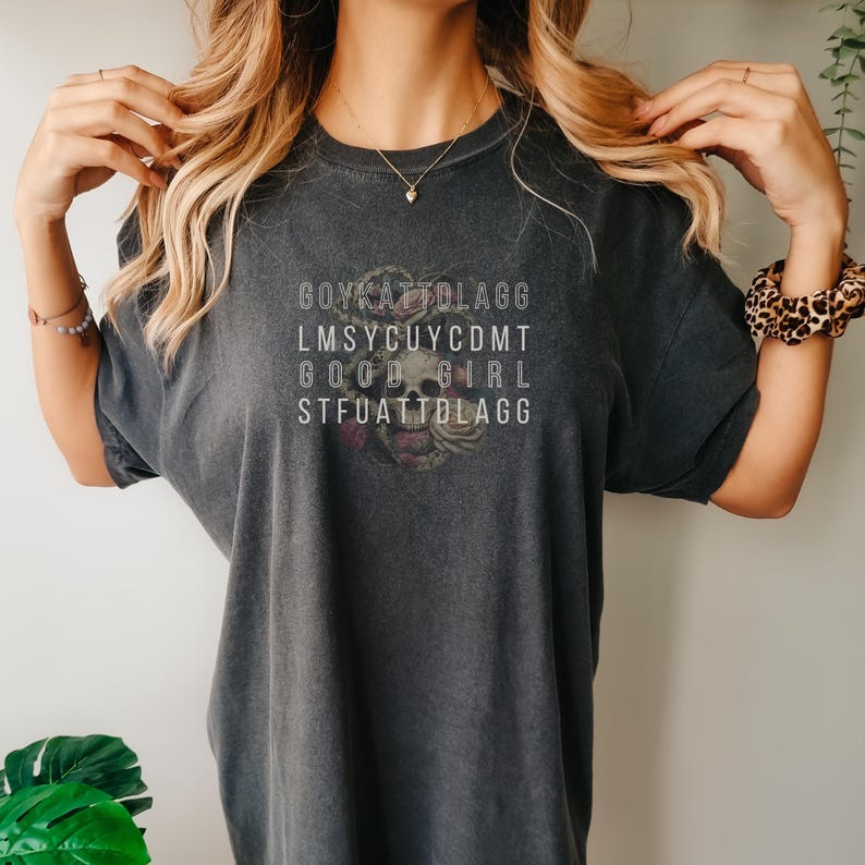 May include: A dark grey t-shirt with a skull and floral design. The text on the shirt reads "GOYKATDLAGG LMSYCUYCDMT GOOD GIRL STFUATTDLAGG".