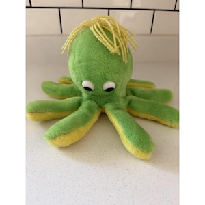 May include: A plush octopus toy featuring a bright green top and yellow underside. The creature has eight soft tentacles, two large white plastic eyes with black pupils, and a tuft of yellow yarn hair attached to the top of its rounded head.