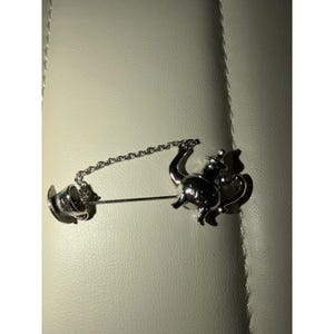 Könnte beinhalten: Eine Brosche aus silberfarbenem Metall, die eine Miniatur-Teekanne und eine passende Teetasse zeigt, welche durch eine feine Gliederkette verbunden sind. Die Teekanne befindet sich rechts an der Nadel, während die Tasse links an der Kette hängt.