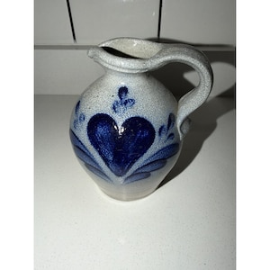 May include: A salt-glazed stoneware pitcher with a speckled light grey finish and a prominent dark blue hand-painted heart motif on the front. The vessel features a curved handle, a wide pouring spout, and a rounded body with a textured surface.