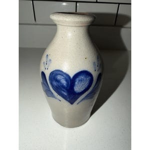 Puede incluir: Un jarrón de gres beige con cuello estrecho y borde acampanado. El cuerpo del jarrón presenta un diseño de corazón azul cobalto pintado a mano en el centro, flanqueado por motivos de hojas azules y pequeños puntos decorativos sobre el corazón.