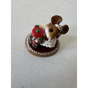Pode incluir: Uma pequena estatueta apresentando um rato castanho com um vestido branco e uma coroa prateada, ao lado de um soldado quebra-nozes em miniatura com chapéu e uniforme vermelhos. As figuras estão montadas numa base redonda com borda branca.