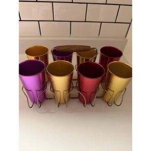 May include: A set of eight metallic tumblers in shades of gold, purple, and red, arranged in a wire rack with a wooden handle. The cylindrical cups feature a smooth, reflective finish and are held securely in the metal frame for easy transport.