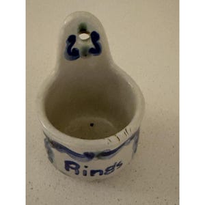 May include: A white ceramic wall-mounted holder with a rounded basin and a back tab featuring a hole for hanging. The exterior is decorated with blue painted scrollwork and the word Rings written in blue script on the front surface.