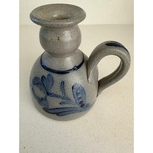 Puede incluir: Un candelabro de gres gris con textura moteada, que presenta una base redondeada, una abertura superior ensanchada y un asa lateral curva. La pieza está decorada con motivos florales de color azul oscuro pintados a mano y una banda azul fina.