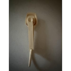 Può includere: Un set di misurini in plastica color crema impilati insieme. Sul manico superiore è chiaramente impresso il testo 1/8 tsp. I misurini presentano una forma a coppa rettangolare e un manico lungo e piatto, tutti collegati alla base.