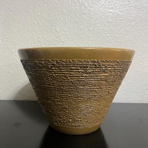 May include: A brown ceramic planter with a textured, ridged exterior. The planter has a wide opening and a slightly flared shape. The rim is a darker shade of brown than the body. The planter is sitting on a black surface.