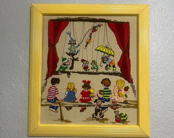 May include: A framed embroidery depicting a puppet show with a cat, monkeys, and turtles.  Children are watching the show from a bench in front of a red curtain.