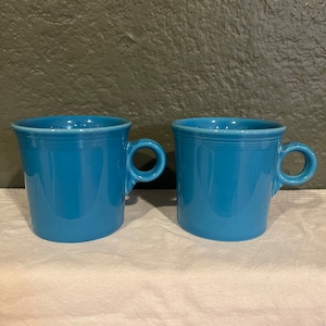 May include: Two blue ceramic mugs with a rounded shape and a single handle. The mugs are sitting on a white surface.