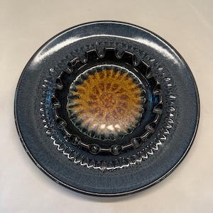 May include: A dark blue ceramic ashtray with a decorative, scalloped edge and a smaller, concentric circle in the center with a brown and yellow glaze.