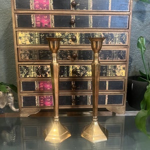 May include: Two gold metal candle holders with a geometric design. The candle holders are sitting on a glass table in front of a wooden cabinet with drawers. The drawers are decorated with book spines.
