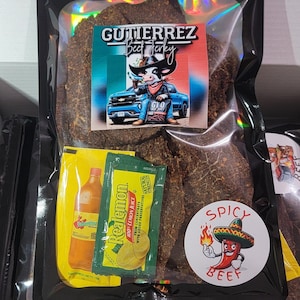 May include: A black package of Gutierrez Beef Jerky with a colorful label featuring a cartoon cow in a cowboy hat. The package includes lemon juice packets and a "Spicy Beef" sticker.