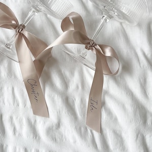 May include: Two champagne flutes with beige satin ribbons tied around the stems. The ribbons are labeled "Isbn" and "Christina".