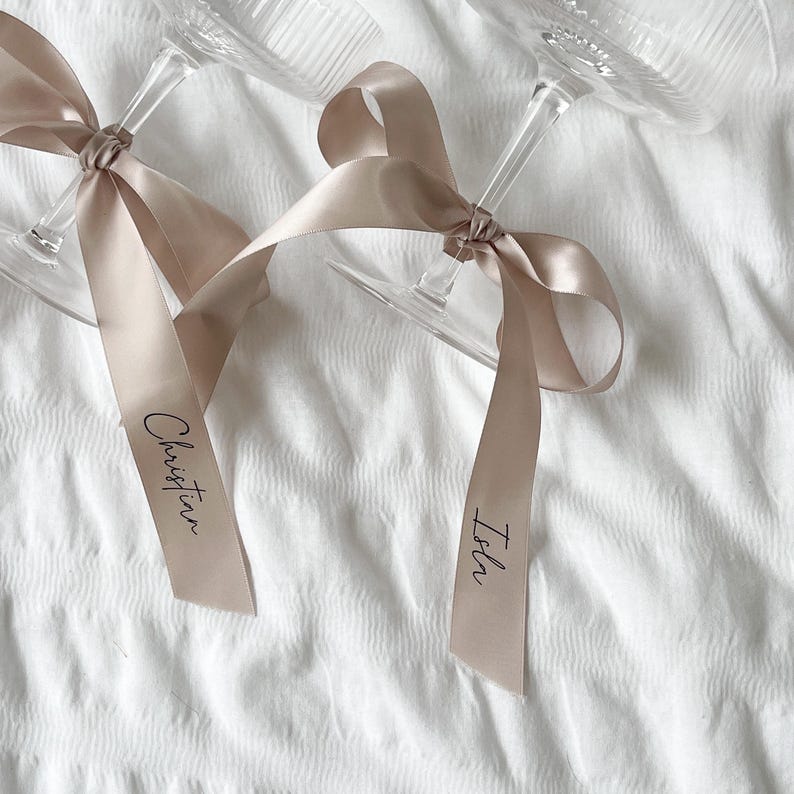 May include: Two champagne flutes with beige satin ribbons tied around the stems. The ribbons are labeled "Isbn" and "Christina".
