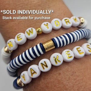 May include: Three bracelets with white beads and gold lettering spelling out "NEW YORK YANKEES". The middle bracelet is a blue and white striped bracelet.