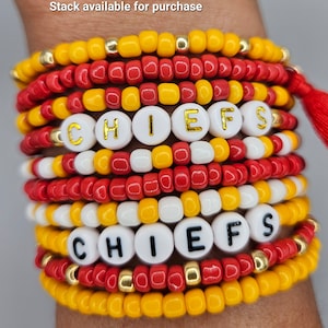 May include: A stack of beaded bracelets in red, yellow, white, and gold. Some bracelets spell out "CHIEFS" with white letter beads. The bracelets are worn on a wrist.