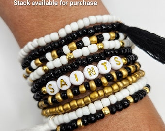 May include: A stack of black, white, and gold beaded bracelets with white beads that spell out "SAINTS" in gold letters. Each bracelet has a black or white tassel.