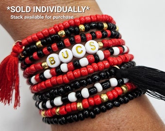 Puede incluir: Una pila de pulseras de cuentas rojas, negras y blancas con detalles dorados. Las pulseras deletrean "Bucs" en letras blancas con contornos dorados. Las pulseras tienen borlas negras.