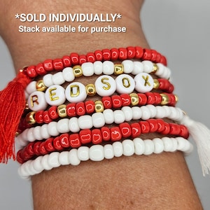 May include: A stack of red and white beaded bracelets with gold beads and white letter beads spelling "RED SOX". The bracelets have red tassels on each end. The text "*SOLD INDIVIDUALLY* Stack available for purchase" is above the bracelets.