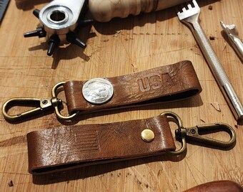 May include: Two brown leather keychains with metal clasps. One keychain has a silver coin with a buffalo design and the word "USA" embossed on the leather. The other keychain has a small American flag design embossed on the leather.
