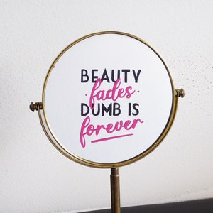 May include: A gold-colored vanity mirror with a round, white surface that reads "BEAUTY fades. DUMB IS forever".